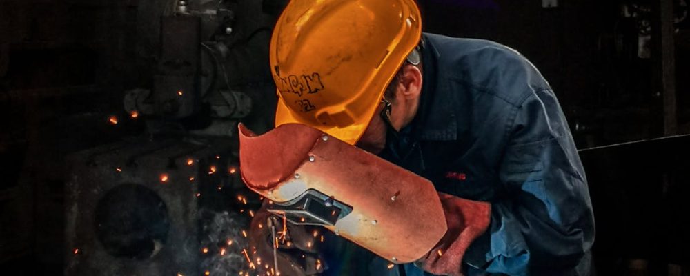 A welder wearing protective gear works on metal in an industrial workshop, surrounded by sparks.