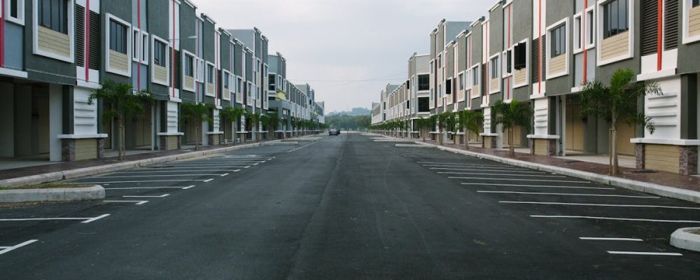 A quiet, empty urban street flanked by modern condominiums under a cloudy sky, with ample parking space.